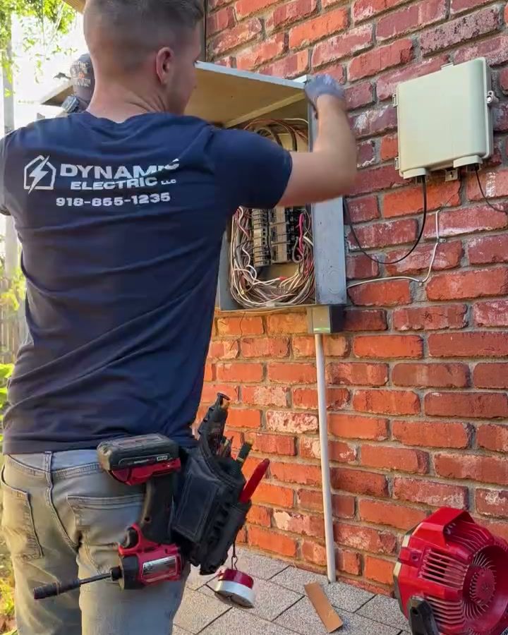 An electrician from behind in a navy Dynamic Electric shirt and tool belt, taking the cover off an old panel on a brick wall.